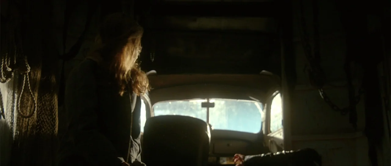 Jeepers Creepers 3 (2017) movie still: medium — The interior of a dark, vintage vehicle with heavy chains hanging on the side walls. A woman sits in…