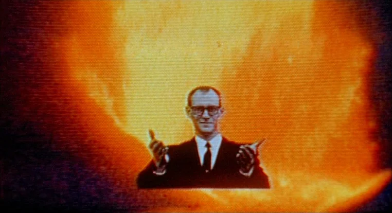 Schizopolis movie still: medium — A man in a suit stands centered against an intense, large orange fire-like glow. The image has heavy analog scan…
