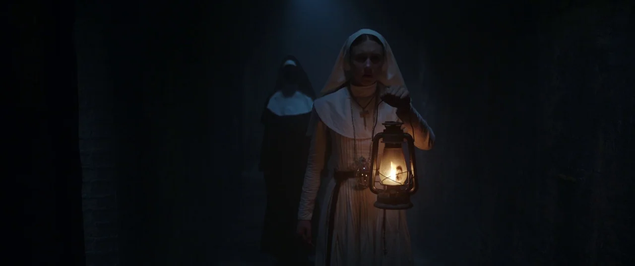 The Nun (2018) movie still: medium — A dark stone passageway with textured walls is enveloped in low light and fog. A young nun in white carries a…