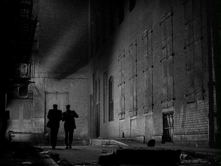 Killer’s Kiss (1955) movie still: wide — A dark city alleyway features a tall brick building with boarded-up windows and doors. Two men in dark…