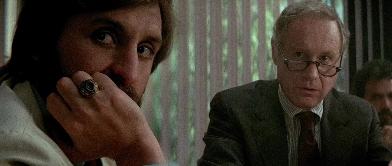 The Entity (1982) movie still: medium — A dimly lit conference room features vertical blinds covering a bright window background. Two men sit at a…