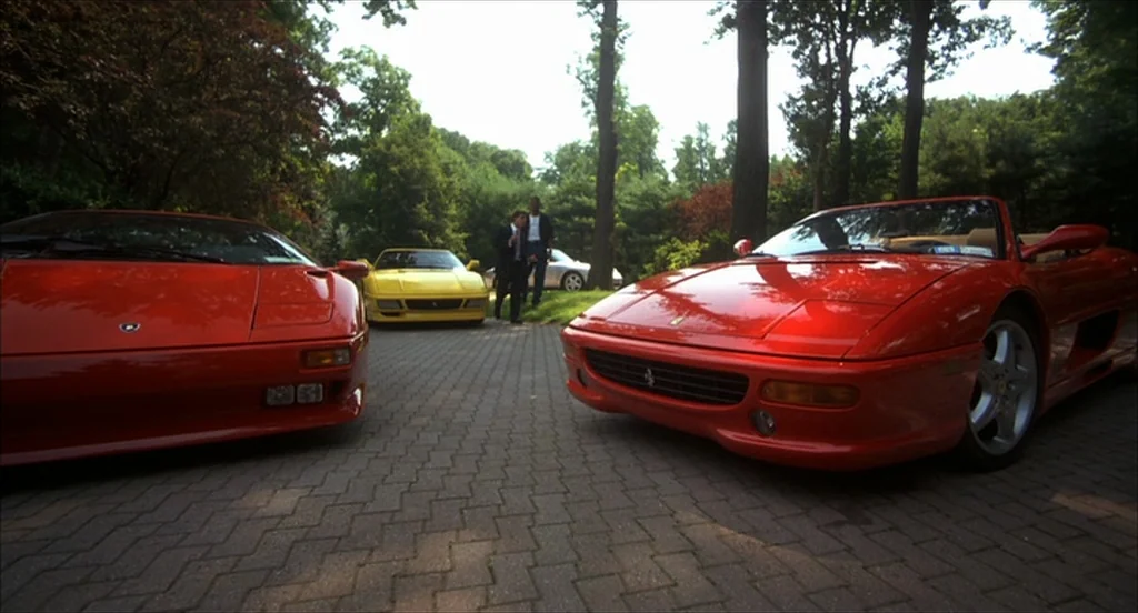 He Got Game (1998) movie still: wide — A paved residential driveway features several luxury sports cars parked amidst lush green trees. Two men in…