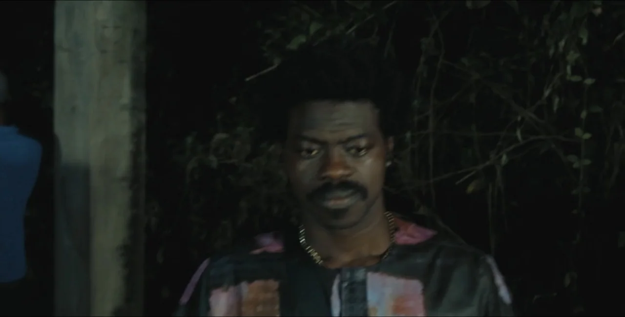 Project Assault (2025) movie still: medium — A dark outdoor environment features a man standing in front of dense foliage. The subject faces the…