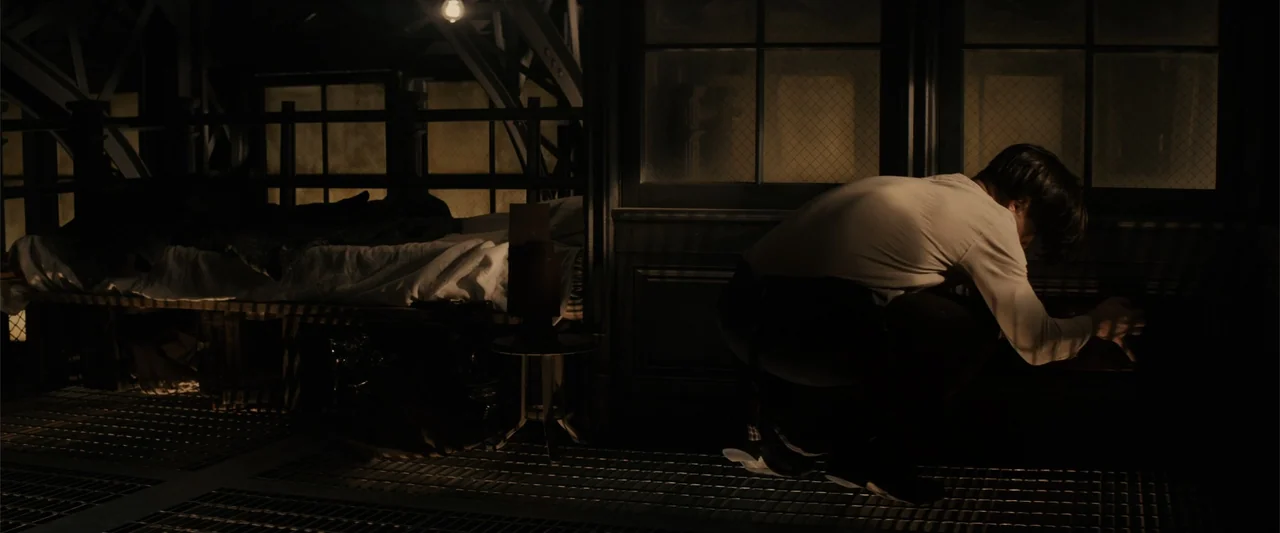 Winter's Tale (2014) movie still: medium — A dimly lit room features a metal floor grate and a wooden bed frame. A man kneels on the floor, facing a…