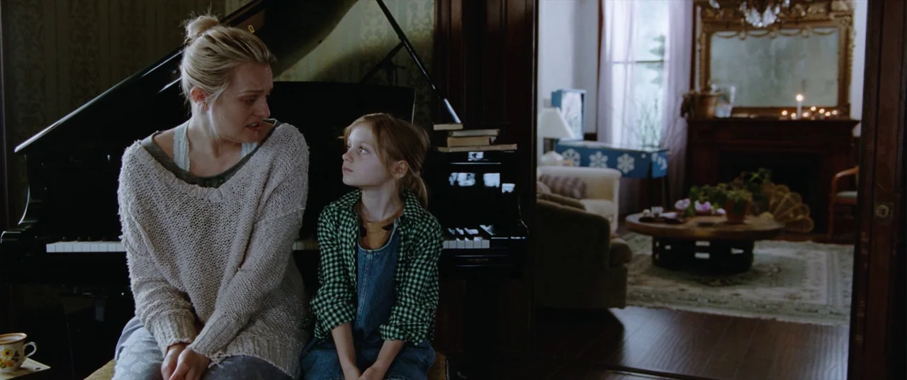 Her Smell (2018) movie still: medium — A woman and young girl sit together in front of a grand piano. Through the doorway, an adjacent room with a…