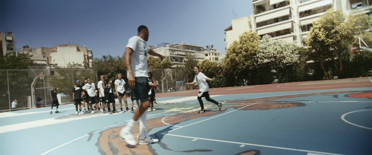 Giannis: The Marvelous Journey (2024) movie still: fullbody — An outdoor painted basketball court sits in an urban environment with multi-story…