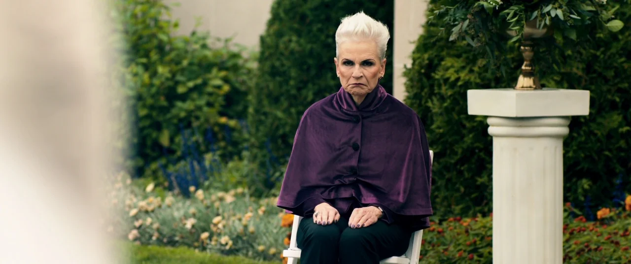 Ready or Not (2019) movie still: medium — An outdoor floral area features an elderly woman sitting on a white chair. She maintains a stern facial…