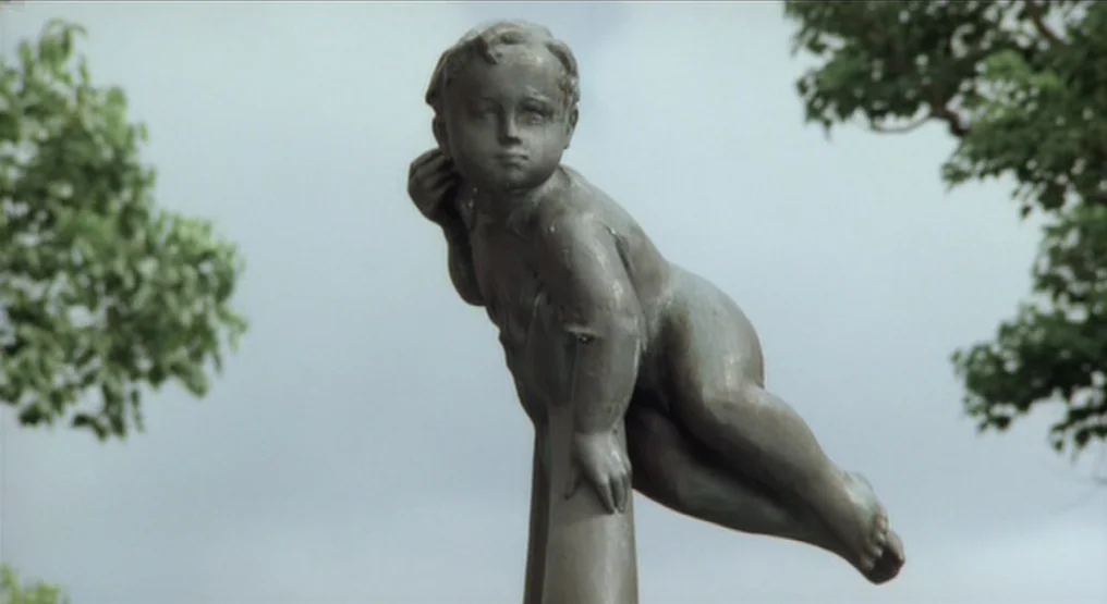 Rhapsody in August (1991) movie still: closeup — A bronze statue of a child rests atop a metal pole in a park. Green foliage and a cloudy sky are…