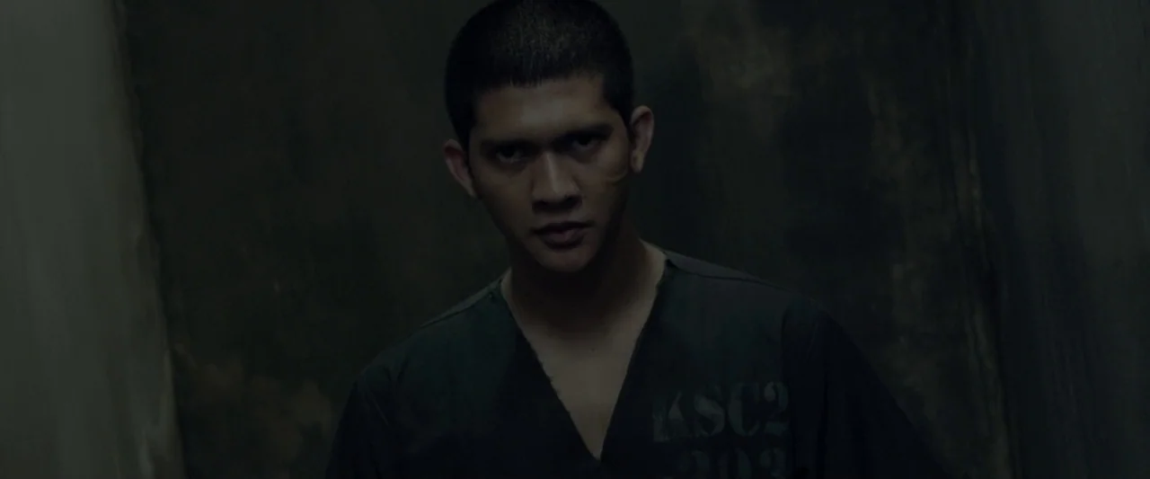 The Raid 2 (2014) movie still: medium — A dimly lit industrial corridor with textured dark walls forms a narrow, confined space. A man in a dark…