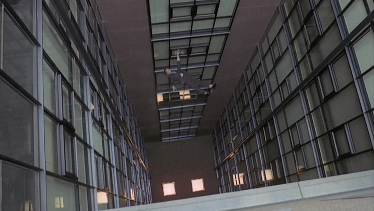 The Black Belly of the Tarantula (1971) movie still: wide — A man falls through a tall glass atrium with repeating window panels on both sides. The…