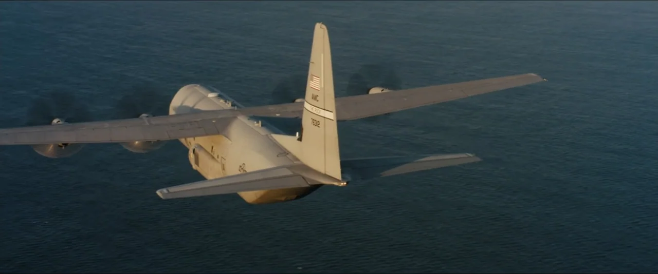 World War Z (2013) movie still: wide — A military C-130 Hercules transport aircraft flies at low altitude over open water. The airplane is viewed…