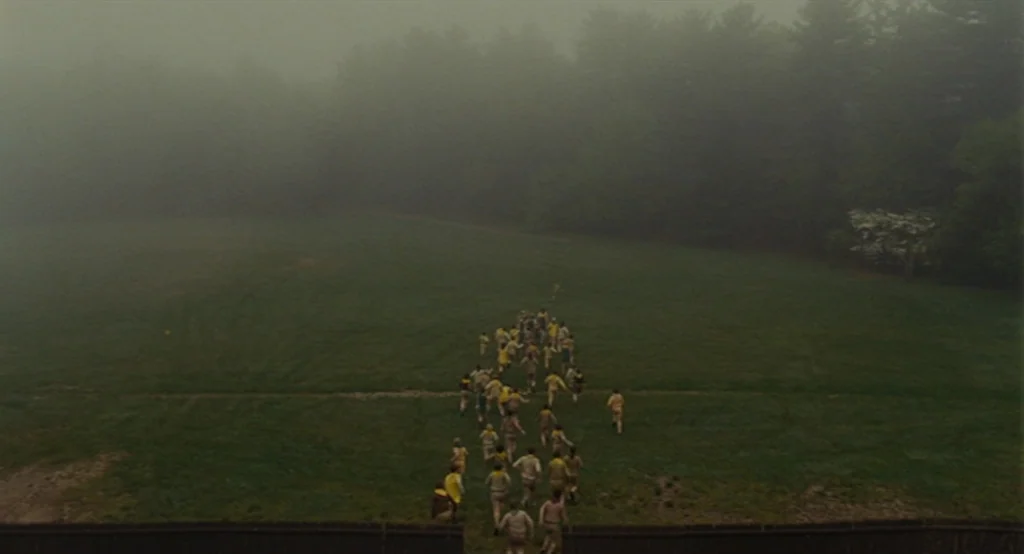 Moonrise Kingdom (2012) movie still: wide — A wide shot shows a group of children running across a dense grassy meadow. Thick fog partially obscures…