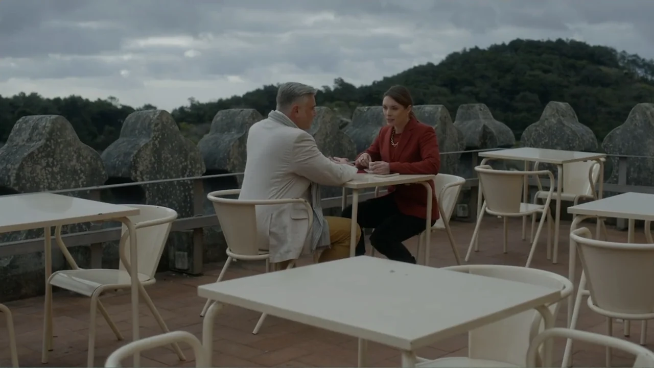 A Pinch of Portugal (2023) movie still: medium — An outdoor terrace with stone battlements overlooks a distant green, cloudy hillside. A man and a…