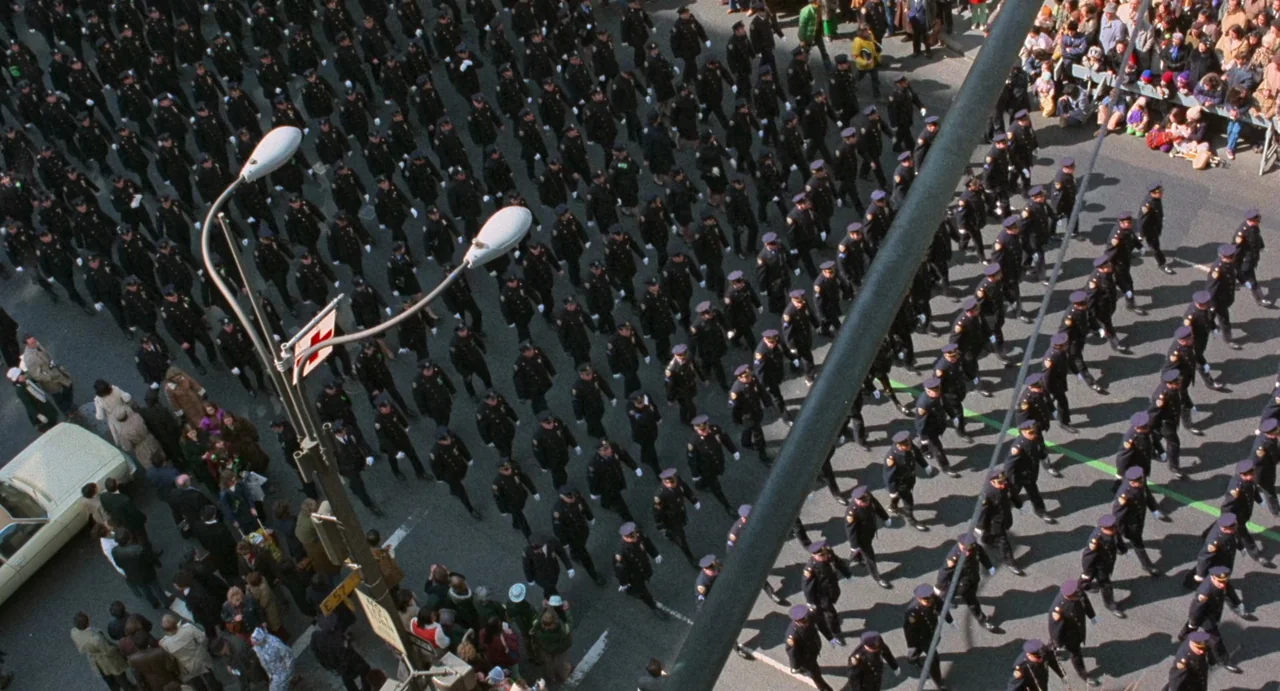 God Told Me To (1976) movie still: wide — A dense formation of uniformed police officers marches across a wide urban thoroughfare. Bystanders gather…