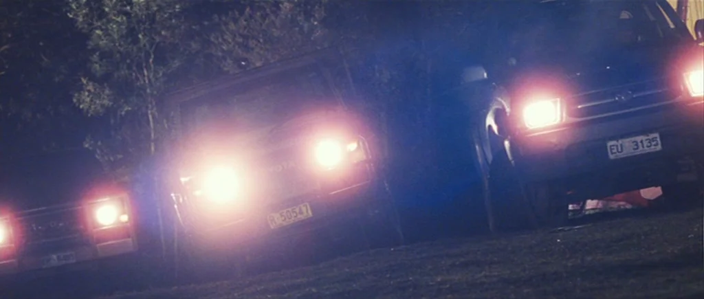 The Hunter (2011) movie still: wide — Three dark off-road vehicles sit on a grassy dirt area at night. Bright, piercing headlights point directly…