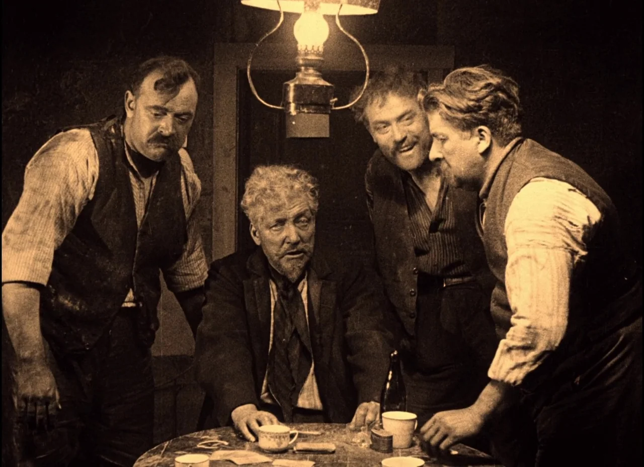 The Phantom Carriage (1921) movie still: medium — Four men in vintage clothing gather around a wooden table in a dimly lit space. An oil lamp hangs…