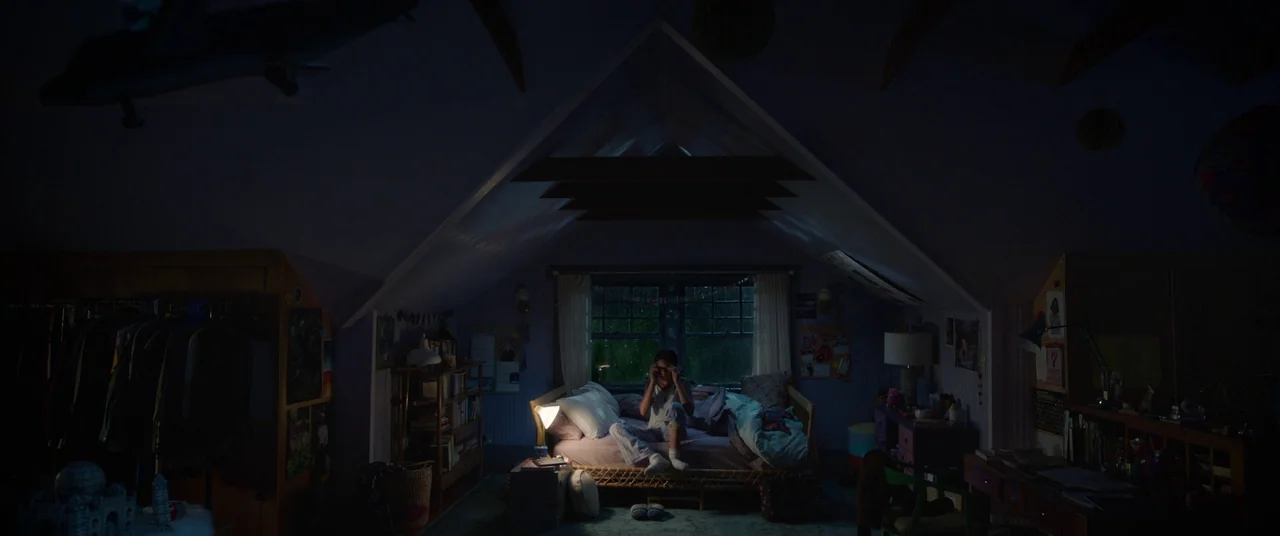 A Wrinkle In Time (2018) movie still: wide — An attic bedroom with sloping walls and dark corners contains a single bed. A lone girl sits on the bed…