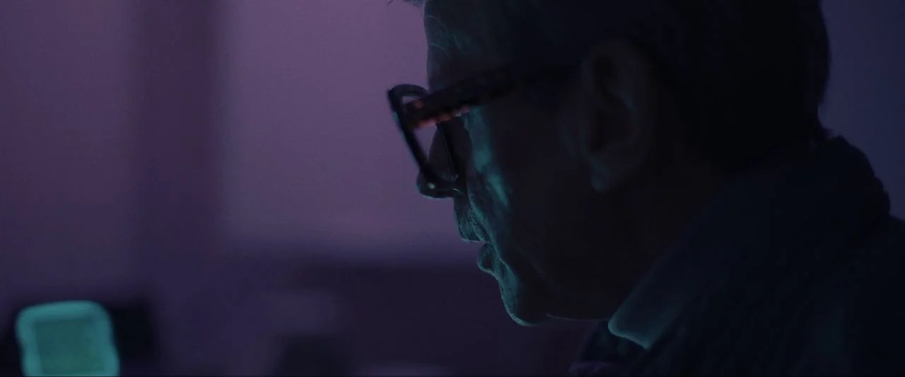 Come True (2020) movie still: closeup — A dimly lit room is bathed in the cool light of a glowing screen. An older man viewed in profile looks…