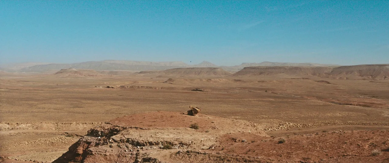 Revenge (2017) movie still: establishing — A vast, arid desert landscape features rolling hills and rocky plateaus under a clear sky. A single small…