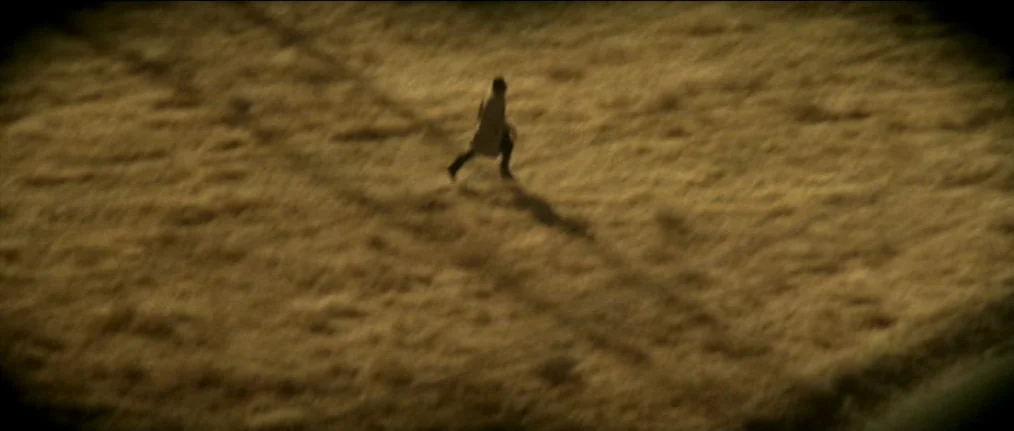 Se7en (1995) movie still: wide — A vast, open field of dry yellow grass stretches under bright, low-angled light. A solitary man in a long coat runs…