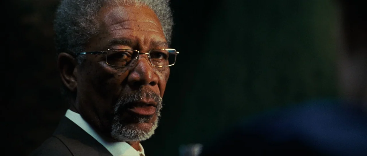 Wanted (2008) movie still: closeup — A man sits in a dimly lit, professional setting with deep shadows behind him. He listens attentively while…