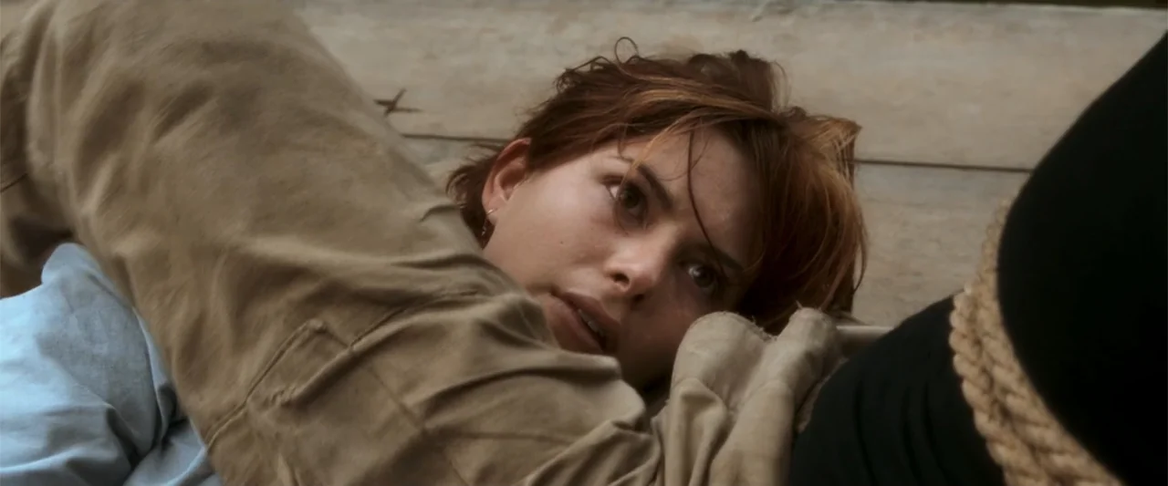 Anaconda (1997) movie still: closeup — A woman lies on a flat, pale ground surface outdoors. She rests on her side while looking towards the viewer.