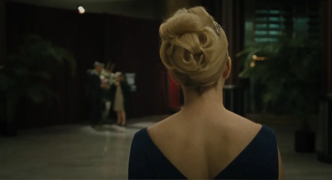Call Jane (2022) movie still: medium — A dimly lit hotel lobby features dark red curtains and blurred potted plants. A woman in a blue dress stands…