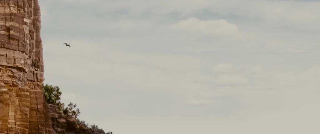 MacGruber (2010) movie still: wide — A tall, layered sedimentary rock cliff stands on the left side of the frame. A lone figure leaps from the edge…