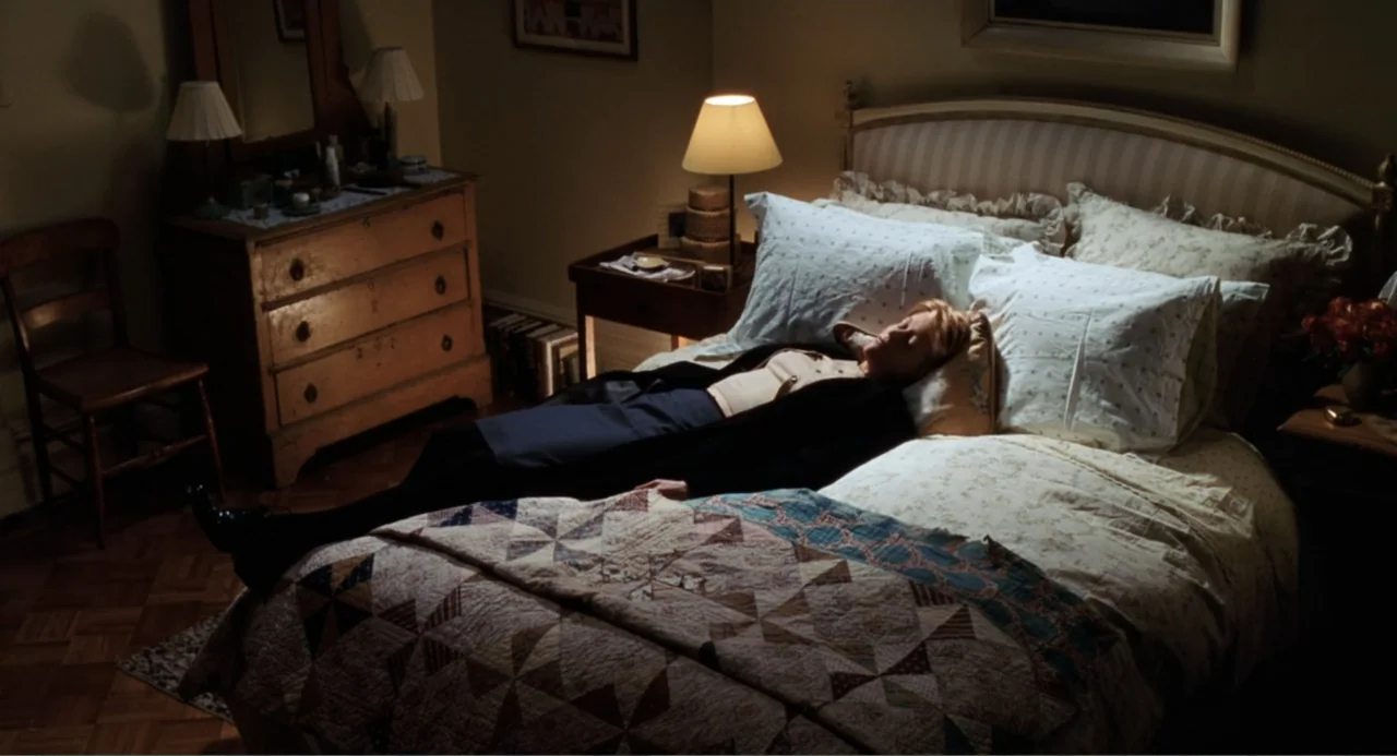 You've Got Mail (1998) movie still: medium — A traditional bedroom contains a wooden dresser, a bedside lamp, and a quilted bedspread. A woman in a…