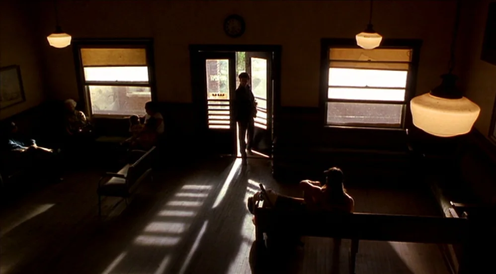 U-Turn (1997) movie still: wide — A sparsely furnished, dark waiting room with wood floors and several wooden benches. A person sits on a bench…