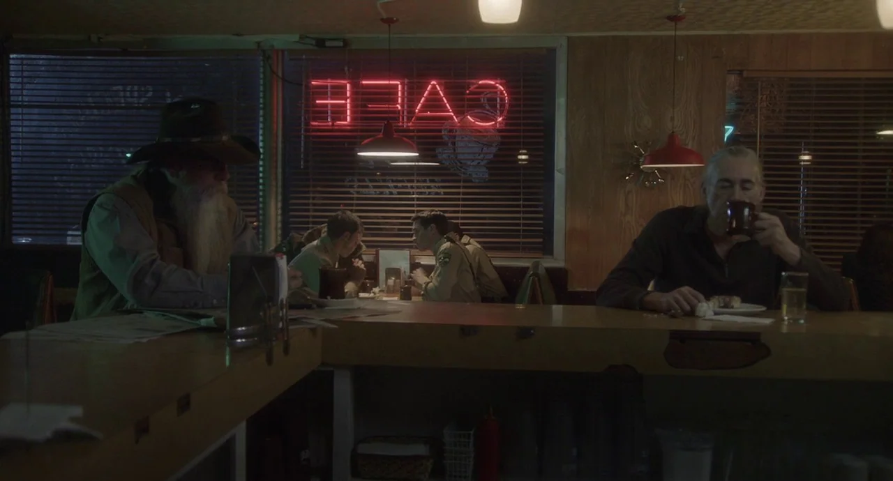 The Wolf of Snow Hollow (2020) movie still: medium — A dimly lit diner interior features wood-paneled walls and a red neon cafe sign. Two men sit at…