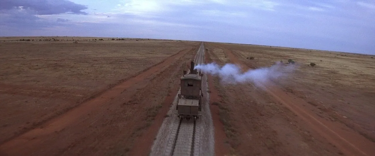 Mad Max Beyond Thunderdome (1985) movie still: wide — A steam locomotive travels along a single track cutting through an arid grassy plain. Red dirt…