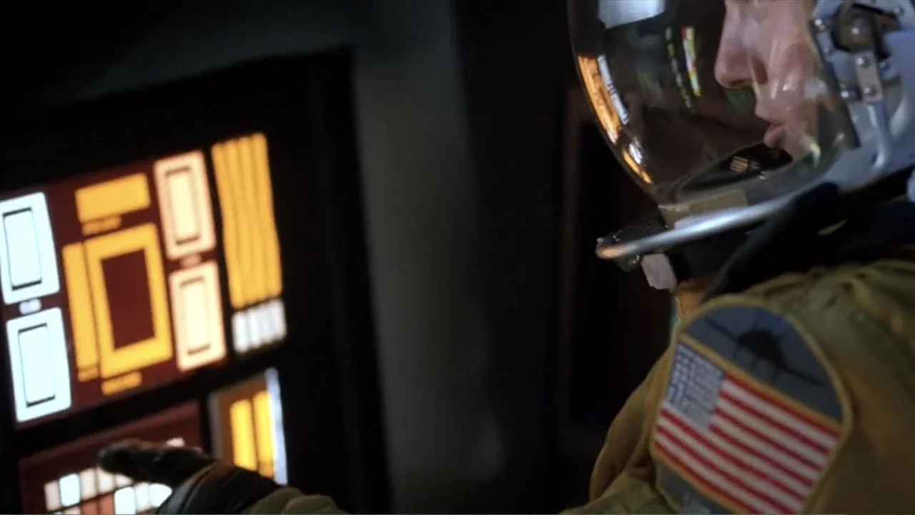 Crisis Point (2012) movie still: closeup — A dark spacecraft interior features a glowing orange instrument panel in the foreground. A pilot in a…