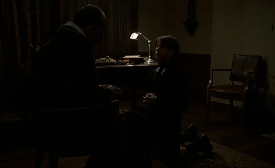 Au Revoir Les Enfants (1987) movie still: medium — A dimly lit, wood-paneled room contains a desk with a lamp and leather chair. A boy kneels on the…