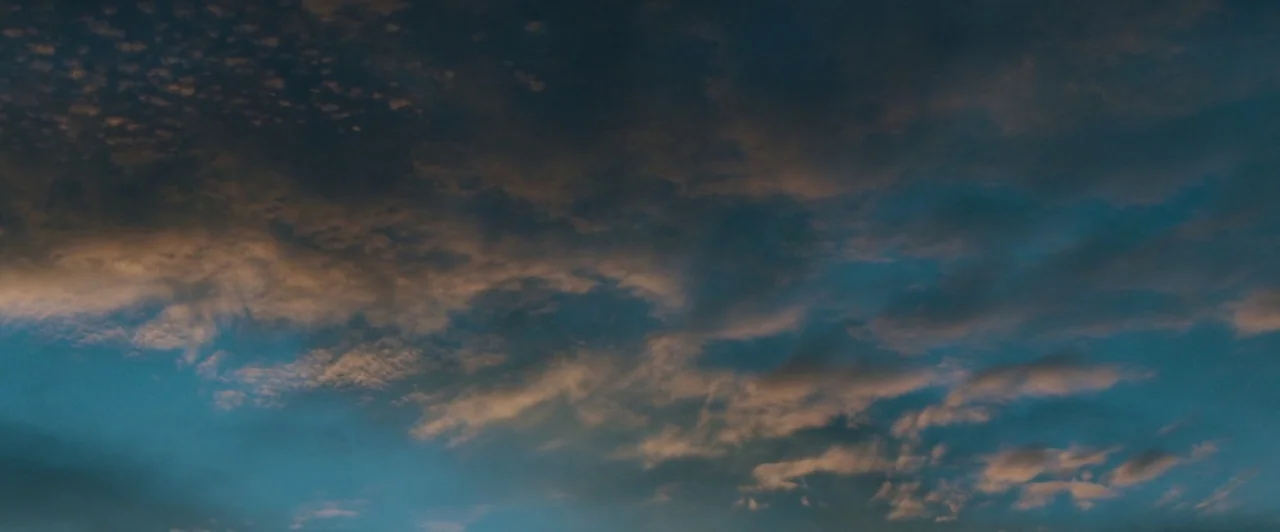 The Long Dumb Road (2018) movie still: establishing — A vast expanse of sky is filled with broken layers of clouds. The late afternoon light casts a…