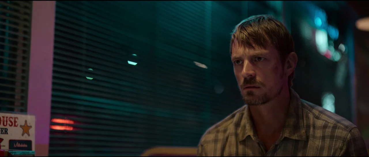 Sympathy For The Devil (2023) movie still: medium — A dark night interior with horizontal window blinds casting thin shadows across the frame. A man…