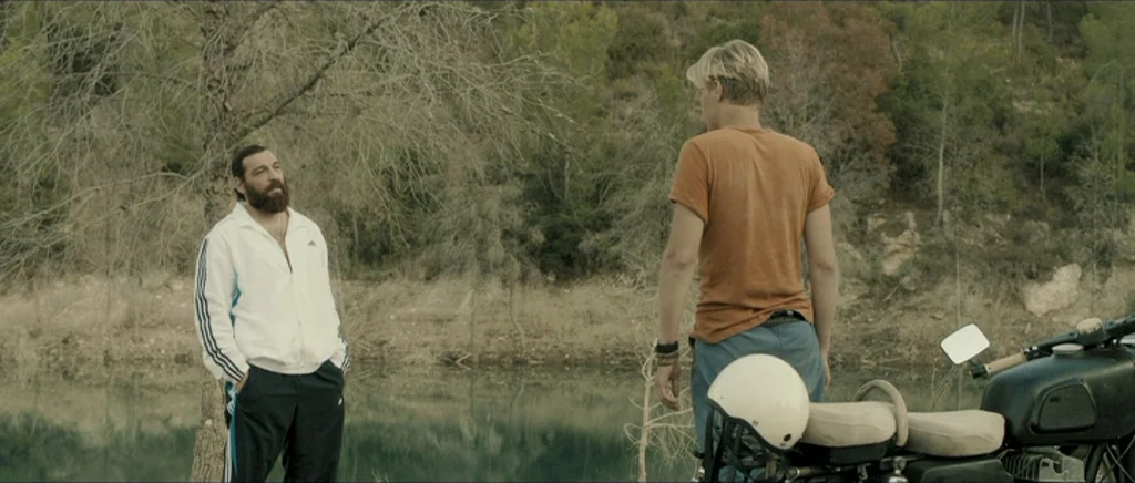 Lasting (Nieulotne) (2013) movie still: fullbody — A quiet lakeshore surrounded by sparse woodland trees with dry brush underfoot. Two men stand in…