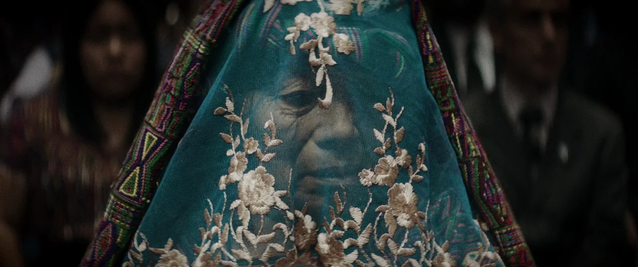 La Llorona (2019) movie still: closeup — An elderly woman sits in a dark, out-of-focus courtroom space. She wears a decorative, embroidered teal…