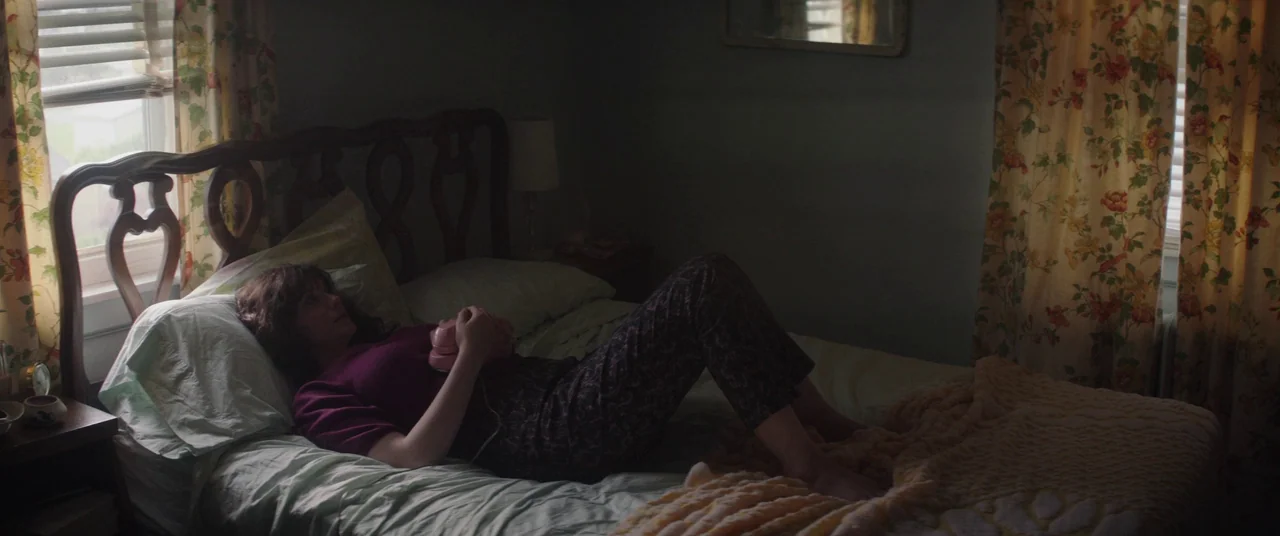 God’s Pocket (2014) movie still: medium — A 1970s style bedroom features floral curtains, a wooden headboard, and faded wall colors. A young woman…