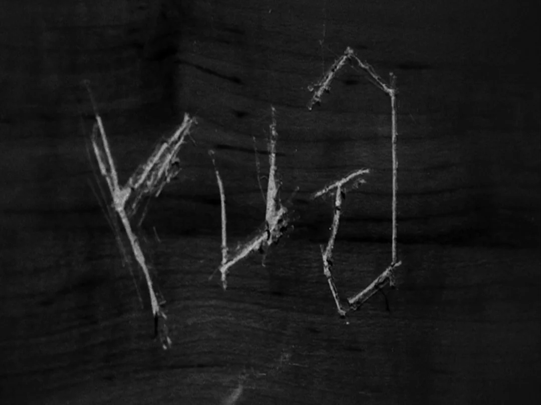 My Winnipeg (2007) movie still: closeup — A dark wooden surface features white carved letters that look like the word YUG. The carving has rough,…