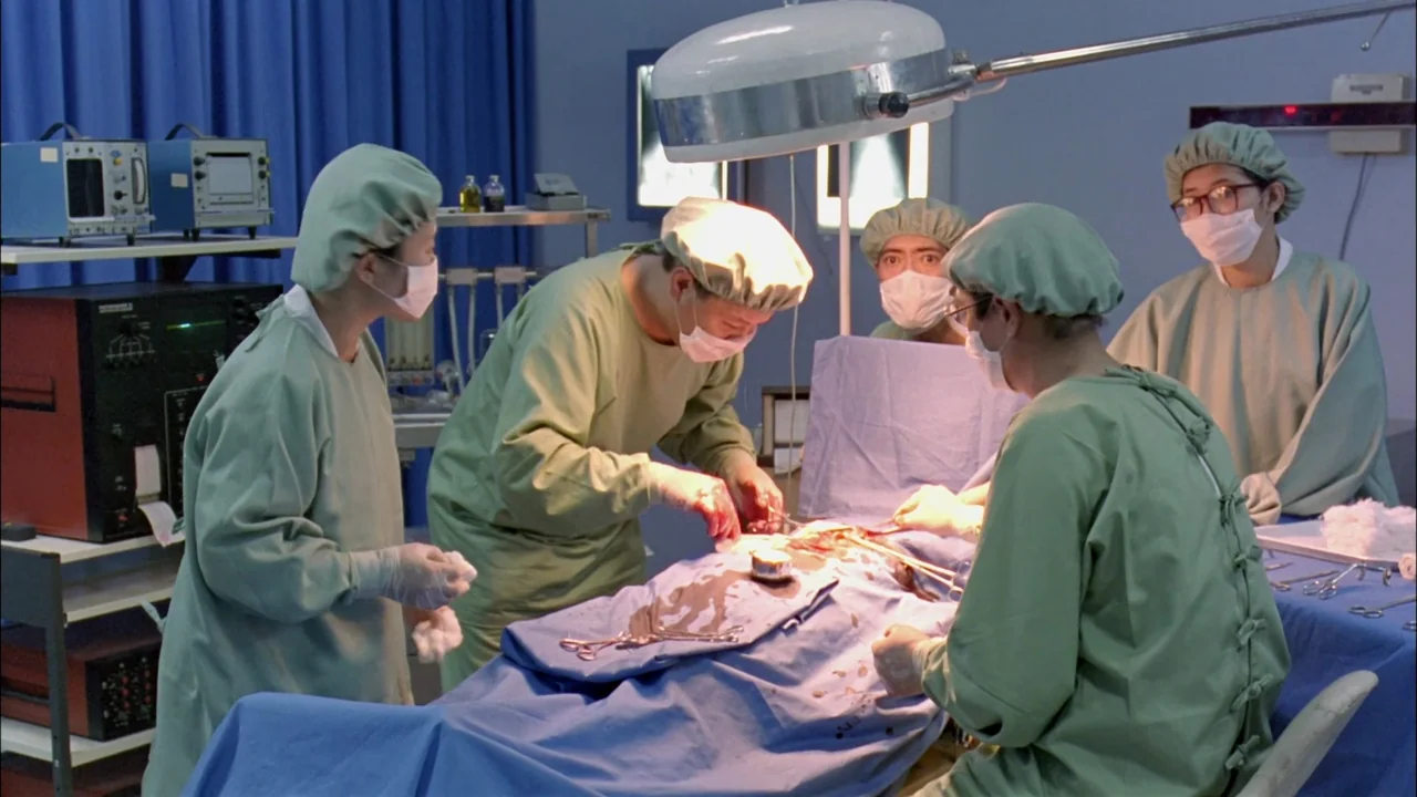 Hard Boiled (1992) movie still: medium — A clinical surgical suite with medical equipment and blue curtains in the background. Several medical staff…