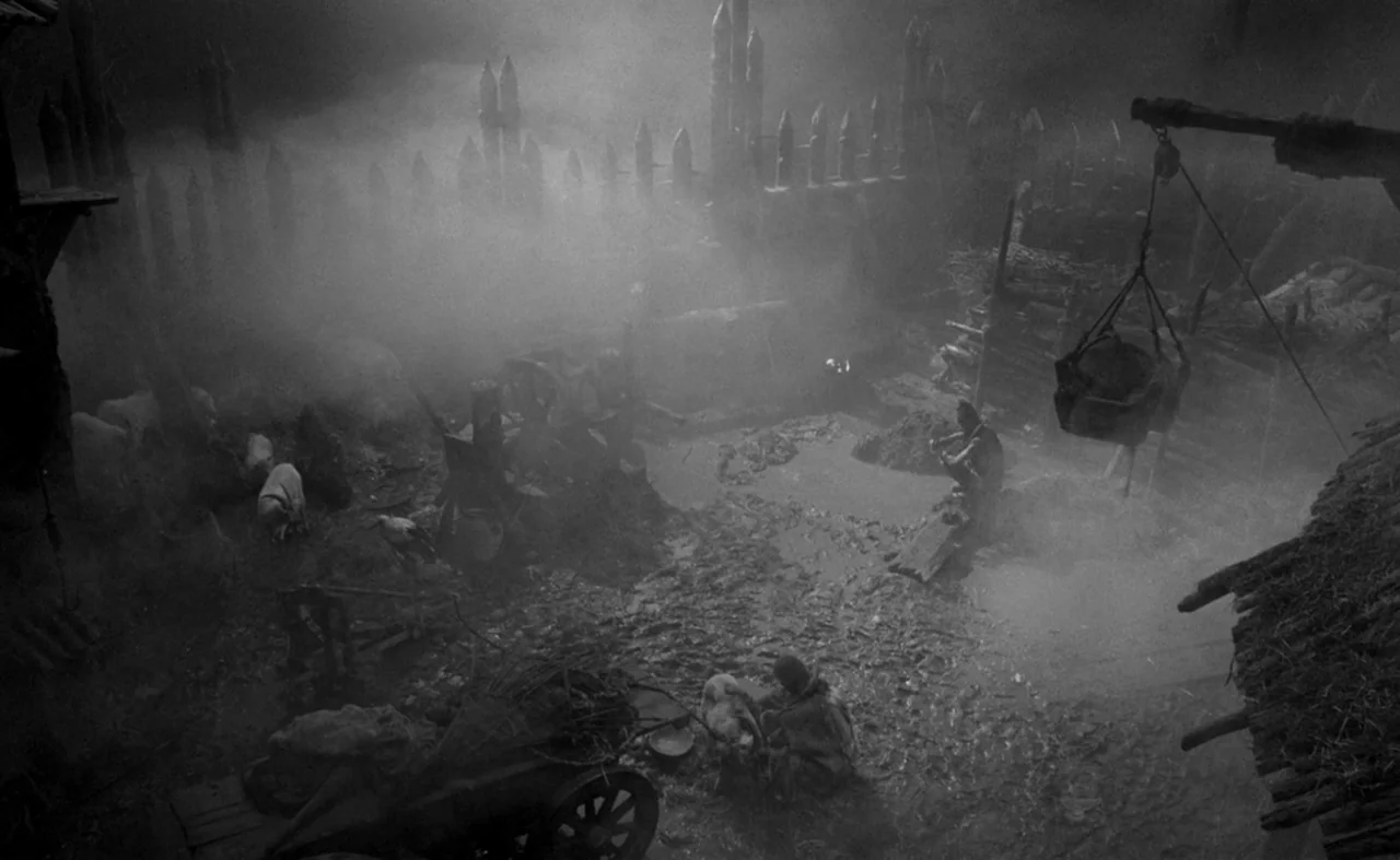 Hard To Be A God (2013) movie still: wide — A dark, mud-filled medieval courtyard is enclosed by a tall, pointed wooden palisade fence. Thick mist…