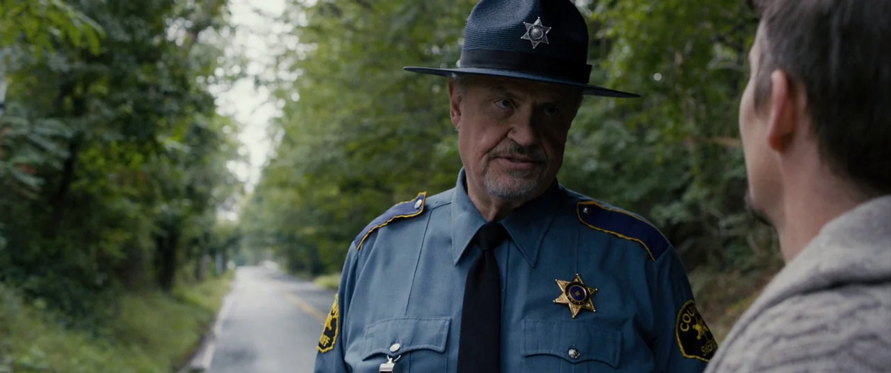 Sinister (2012) movie still: medium — A quiet rural road is flanked by dense green deciduous trees. A county sheriff stands in the foreground…
