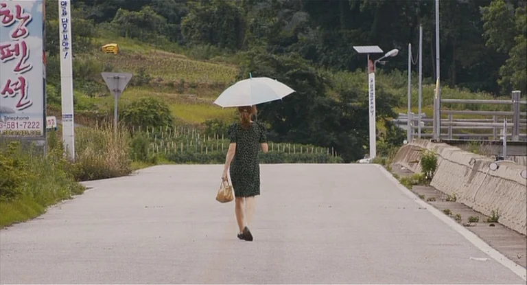 In Another Country (2012) movie still: fullbody — A rural road in South Korea is flanked by verdant hills and a concrete barrier. A woman in a…