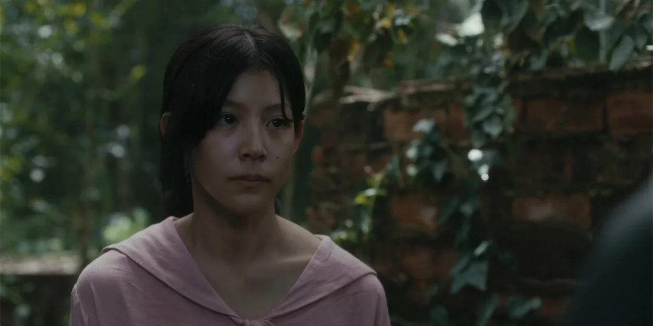 Host (2020) movie still: closeup — A woman stands in a lush outdoor setting with a weathered brick wall nearby. She turns her head slightly as she…