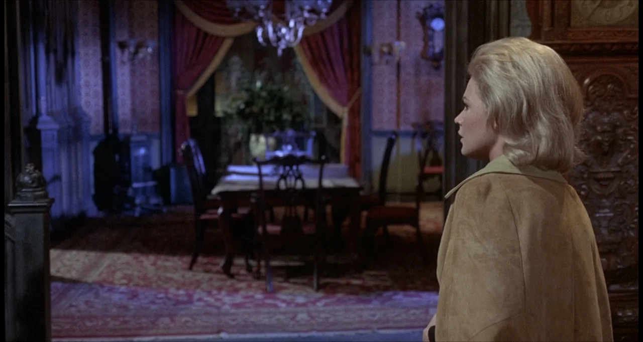 The Dunwich Horror (1970) movie still: medium — A dimly lit room with ornate wooden furniture and heavy red curtains in the back. A blonde woman in…