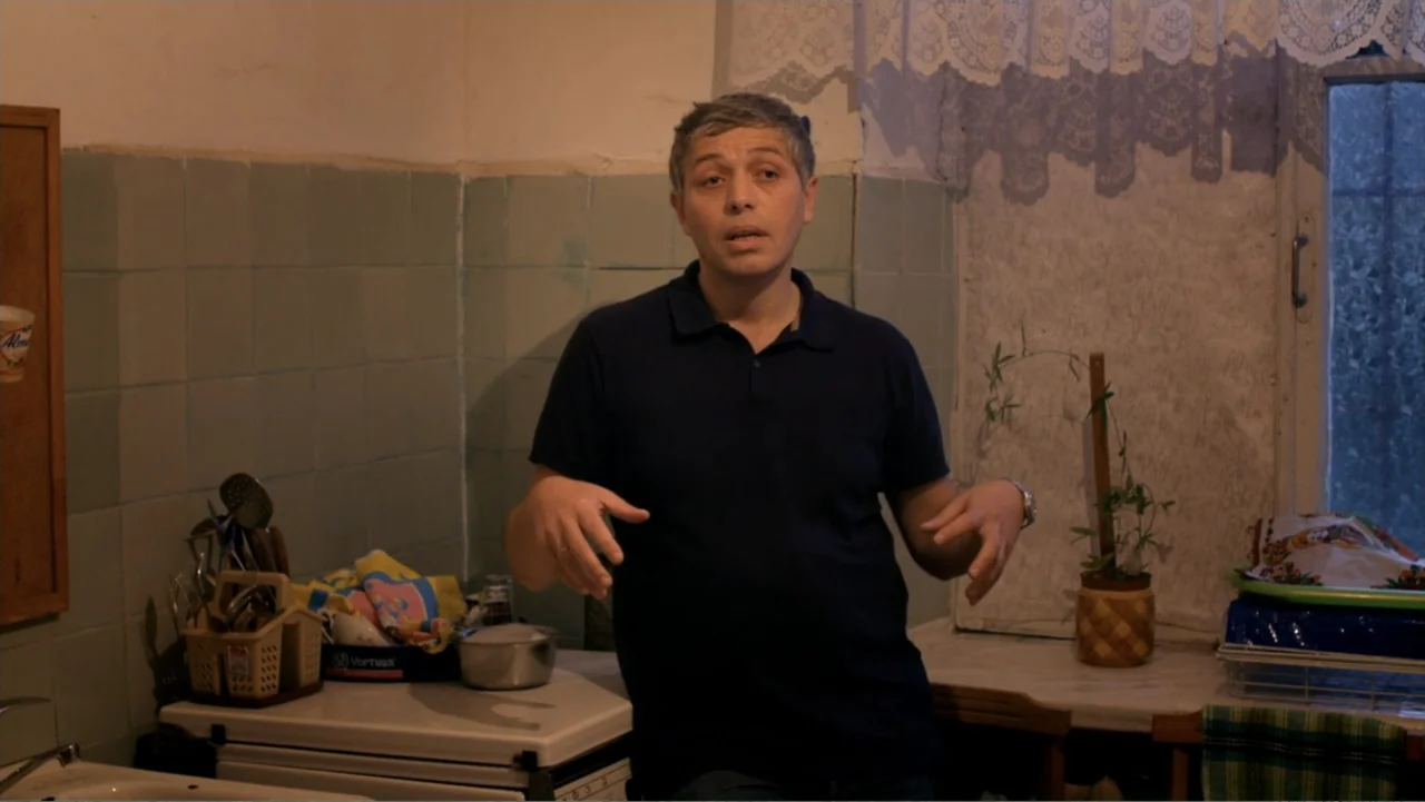 Letters to Max (2014) movie still: medium — A small kitchen features light blue tiled walls and a window with lace curtains. A middle-aged man in a…