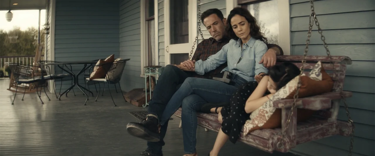 Hypnotic (2023) movie still: medium — A wooden porch with blue horizontal siding features a hanging swing in the foreground. A man and a woman sit…