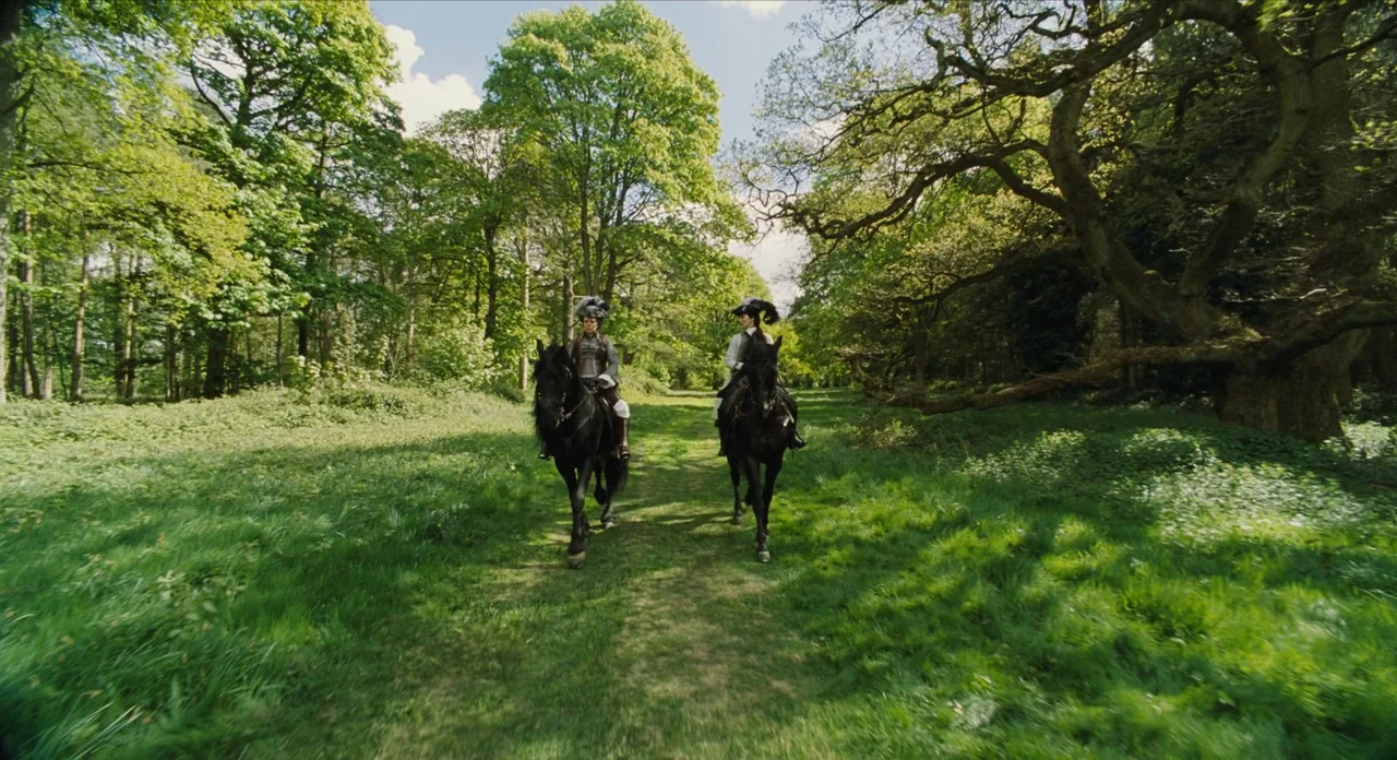 The Favourite (2018) movie still: wide — Two riders on horseback traverse a grassy trail winding through a lush, dense woodland. The path is flanked…
