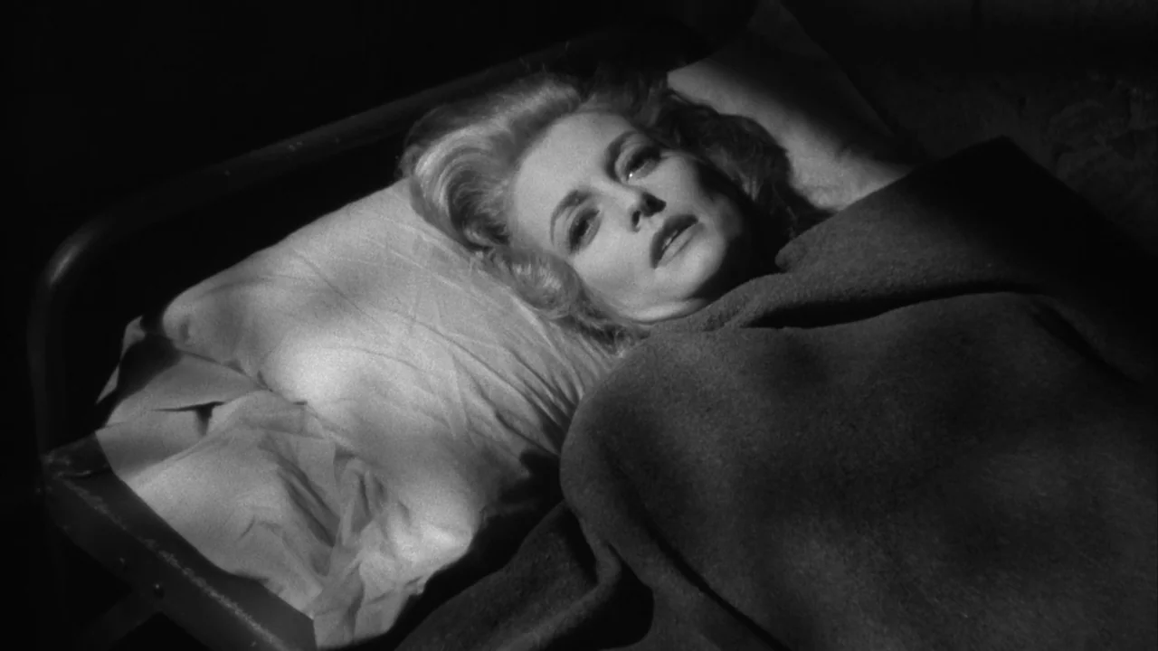 The Naked Kiss (1964) movie still: closeup — A woman lies on a cot with a thin pillow and dark coarse blanket. The space is dark and minimal with…